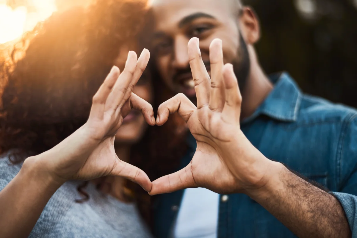 Two people stand close together outdoors, forming a heart shape with their hands in the foreground. their faces are slightly blurred and partially hidden behind their hands.