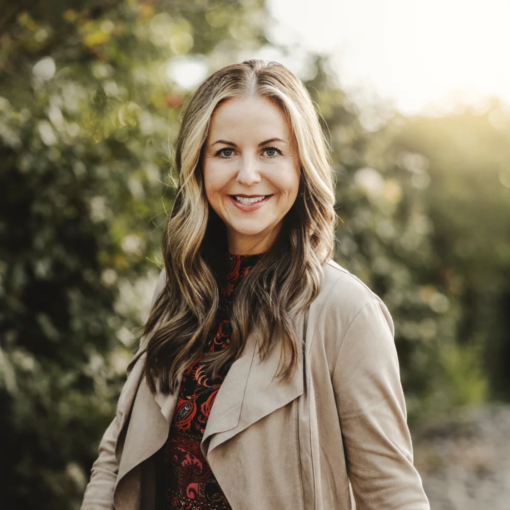A woman with long, wavy hair stands outdoors, smiling at the camera. She is wearing a beige jacket over a patterned top, with greenery in the blurred background.