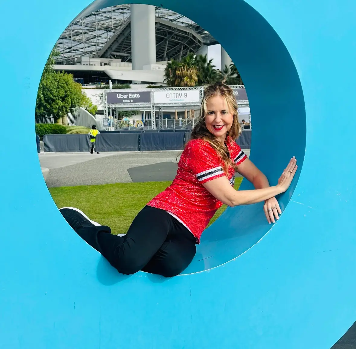 A woman in a red shirt poses inside a large blue circular sculpture outdoors near a stadium entrance.