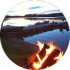 A campfire burns near the edge of a calm lake at sunset, with logs and a dock visible in the background.