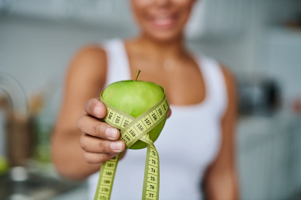 A person in a white tank top holds a green apple wrapped with a measuring tape, suggesting themes of diet or healthy eating.