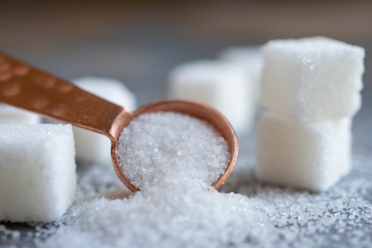 A spoon filled with granulated sugar is surrounded by several white sugar cubes on a gray surface.