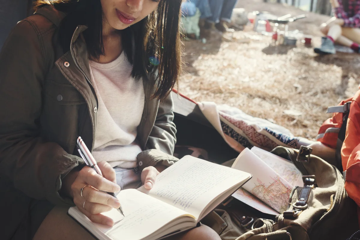 A person sits outdoors writing in a notebook, with an open map and a backpack beside them. other people and trees are visible in the background.