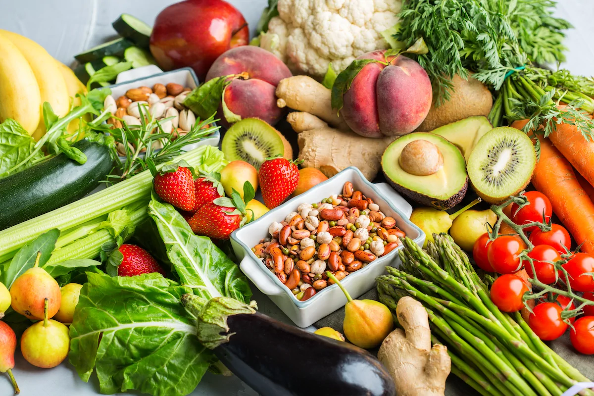An assortment of fresh fruits, vegetables, leafy greens, nuts, and seeds arranged on a table, including strawberries, carrots, avocado, kiwi, tomatoes, and eggplant.